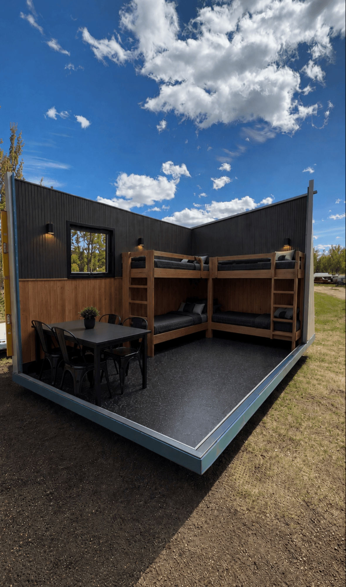 Modern dark ice shack with an open front showing bunk beds and a dining table, under a blue sky.